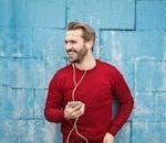 A joyful man in a red sweater smiles while listening to music on his smartphone against a blue brick wall.